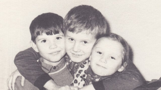 Three children smiling and hugging each other closely, posing for a photo. The image is in black and white, giving it a nostalgic, vintage feel.