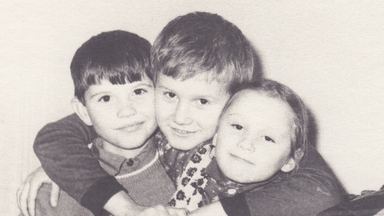 Three children smiling and hugging each other closely, posing for a photo. The image is in black and white, giving it a nostalgic, vintage feel.