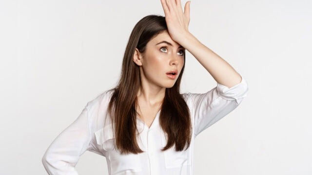 A woman wearing a white shirt slaps her forehead in a facepalm gesture, looking upward with a frustrated expression.