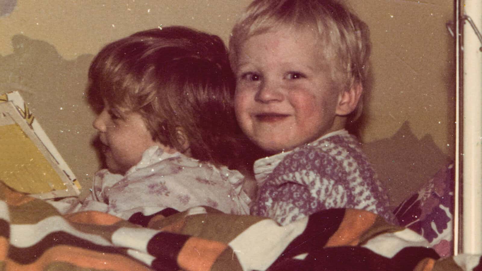 Two young children sitting in bed under a striped blanket, with one child looking toward the camera and the other looking at a book.