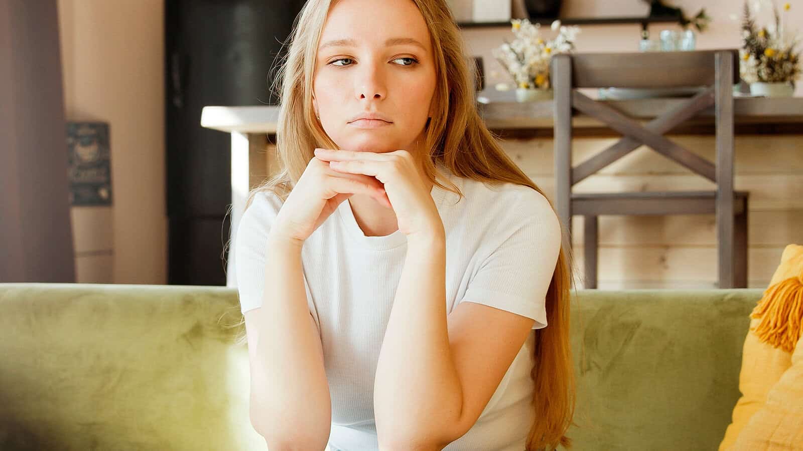 A woman with long hair wearing a white t-shirt rests her chin on her hands while looking to the side.