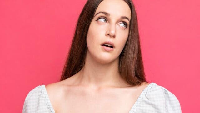 A young woman with long hair looking upward with a confused expression against a pink background.