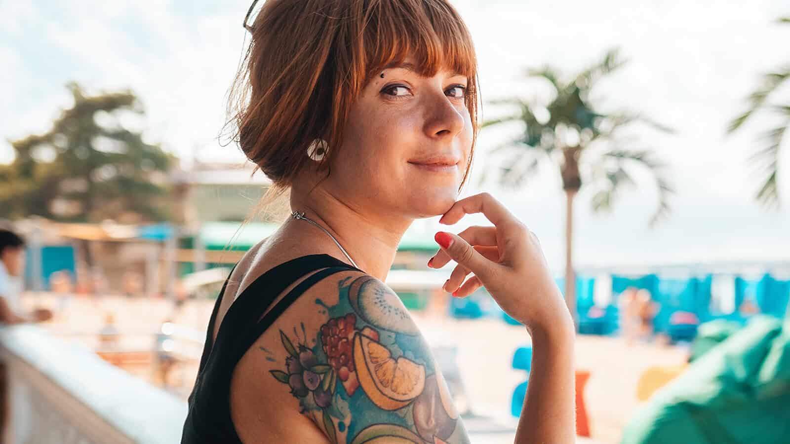 A woman with red hair and a colorful fruit tattoo on her shoulder smiles at the camera, sitting outdoors near a beach with palm trees and blue umbrellas in the background.