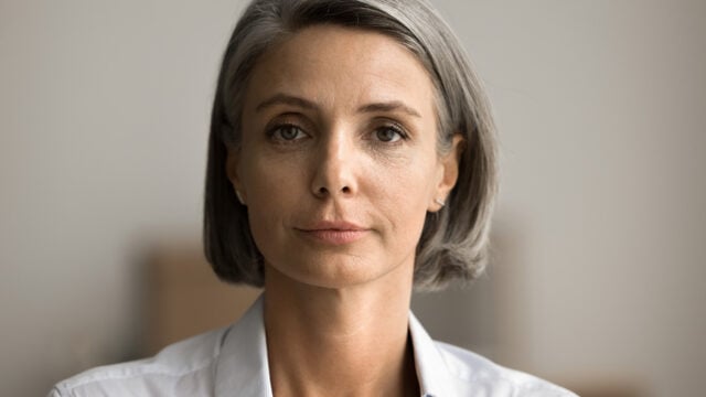 A middle-aged woman with straight gray hair, wearing a light-colored blouse, looks directly at the camera with a neutral expression against a softly blurred background.