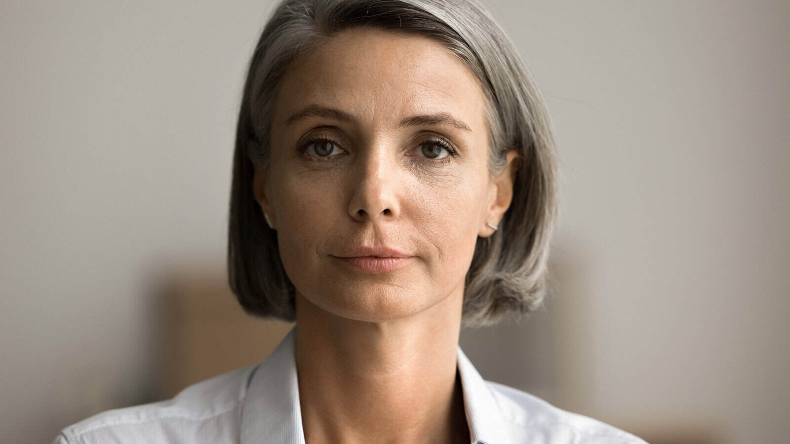 A middle-aged woman with straight gray hair, wearing a light-colored blouse, looks directly at the camera with a neutral expression against a softly blurred background.