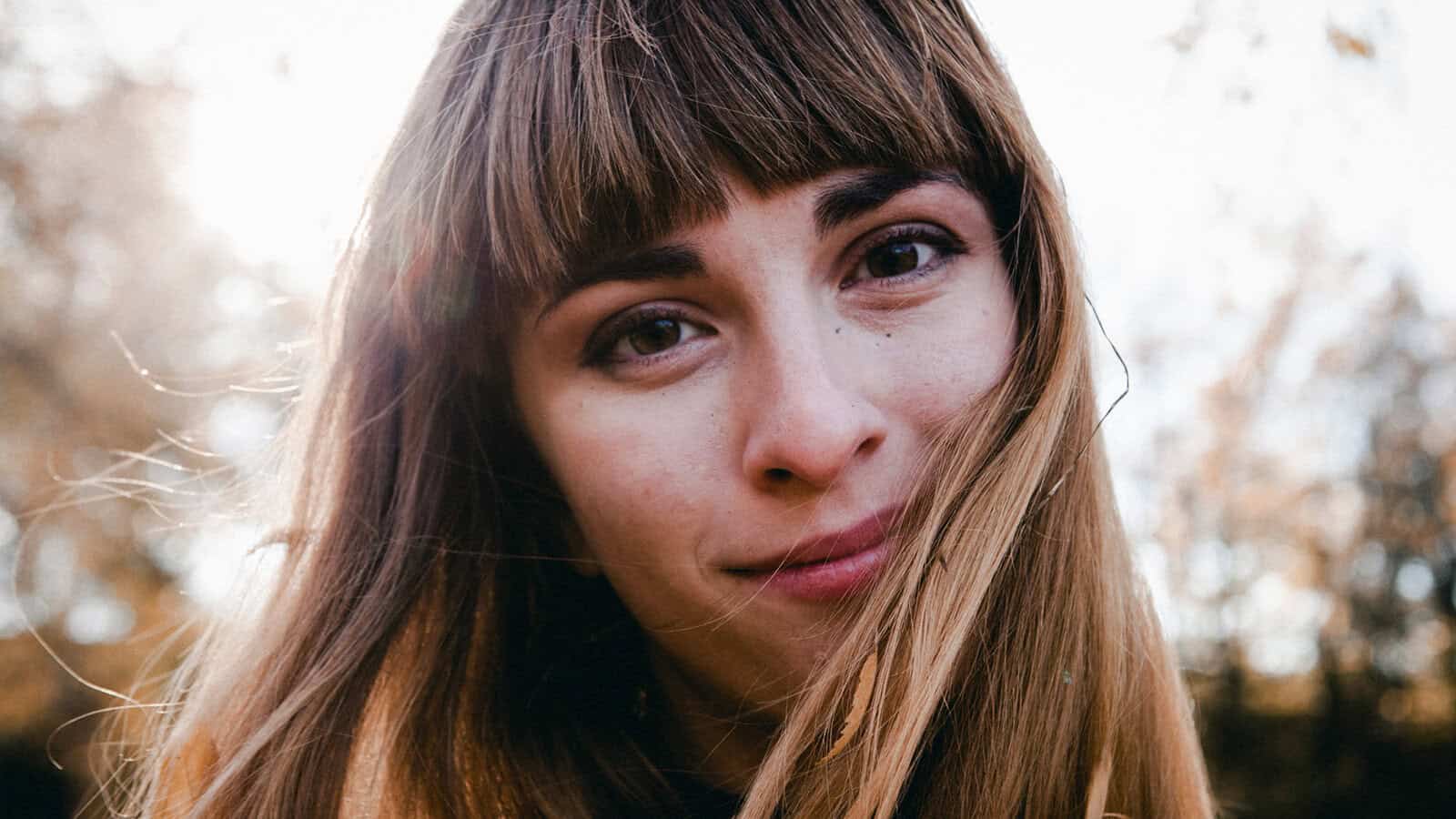 A woman with long brown hair and bangs smiles softly outdoors, sunlight shining behind her, with blurred trees in the background.