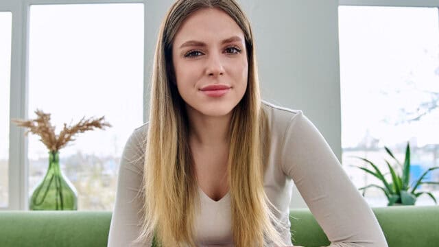 A young woman with straight, long blonde hair sits indoors on a green sofa. She wears a light-colored top and looks directly at the camera. Large windows and houseplants are visible in the background.