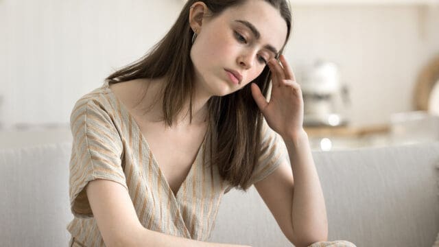 A young woman sitting on a sofa with her hand against her temple, looking down with a concerned expression.