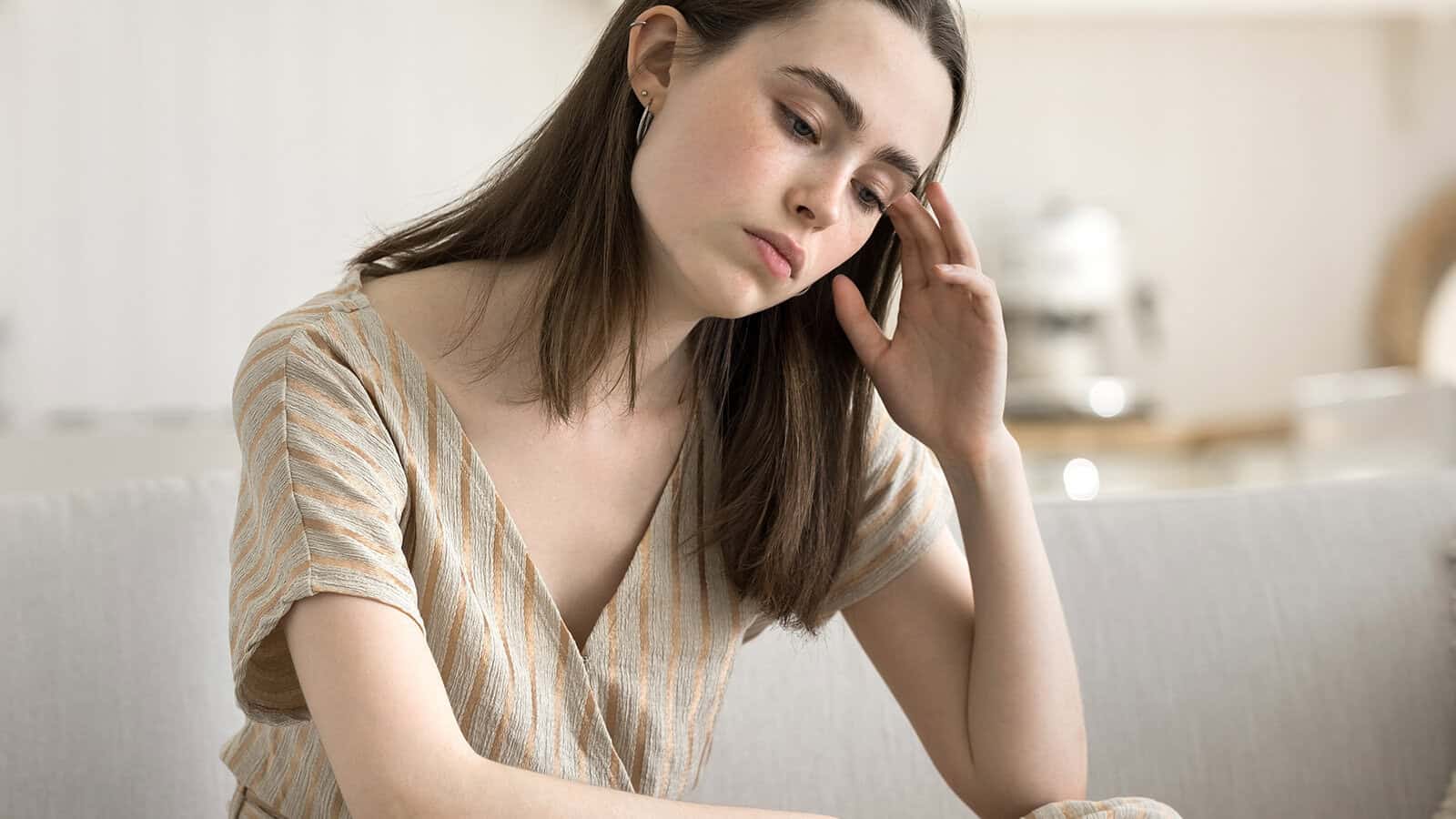 A young woman sitting on a sofa with her hand against her temple, looking down with a concerned expression.