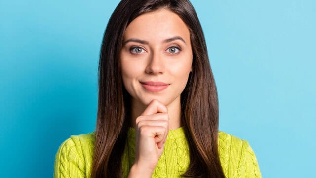 A young woman with long brown hair wearing a lime green sweater rests her chin on her hand against a blue background.