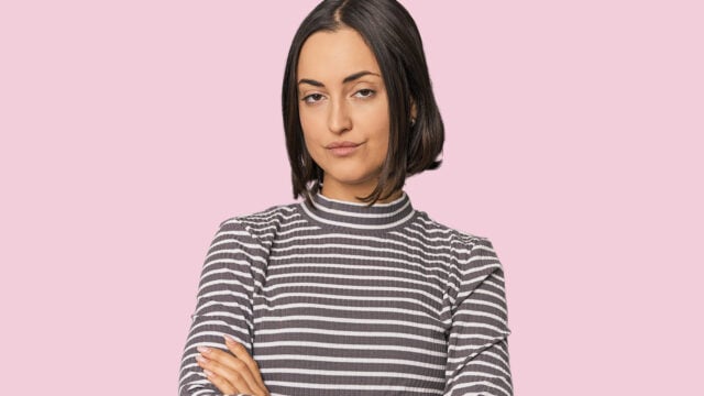 A woman with a bob haircut and a striped shirt stands with her arms crossed against a light pink background.