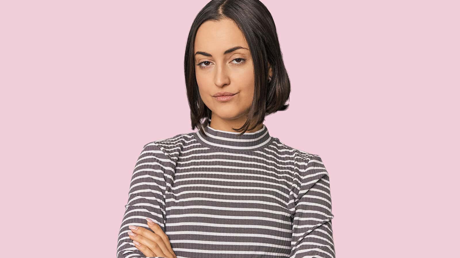 A woman with a bob haircut and a striped shirt stands with her arms crossed against a light pink background.