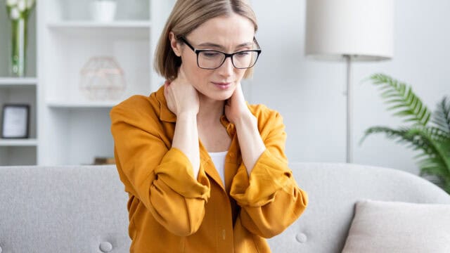 A woman wearing glasses and a yellow shirt sits on a sofa, holding the back of her neck with both hands as if in pain.