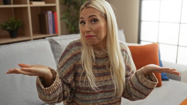 A blonde woman sitting on a sofa shrugs her shoulders with a confused expression.