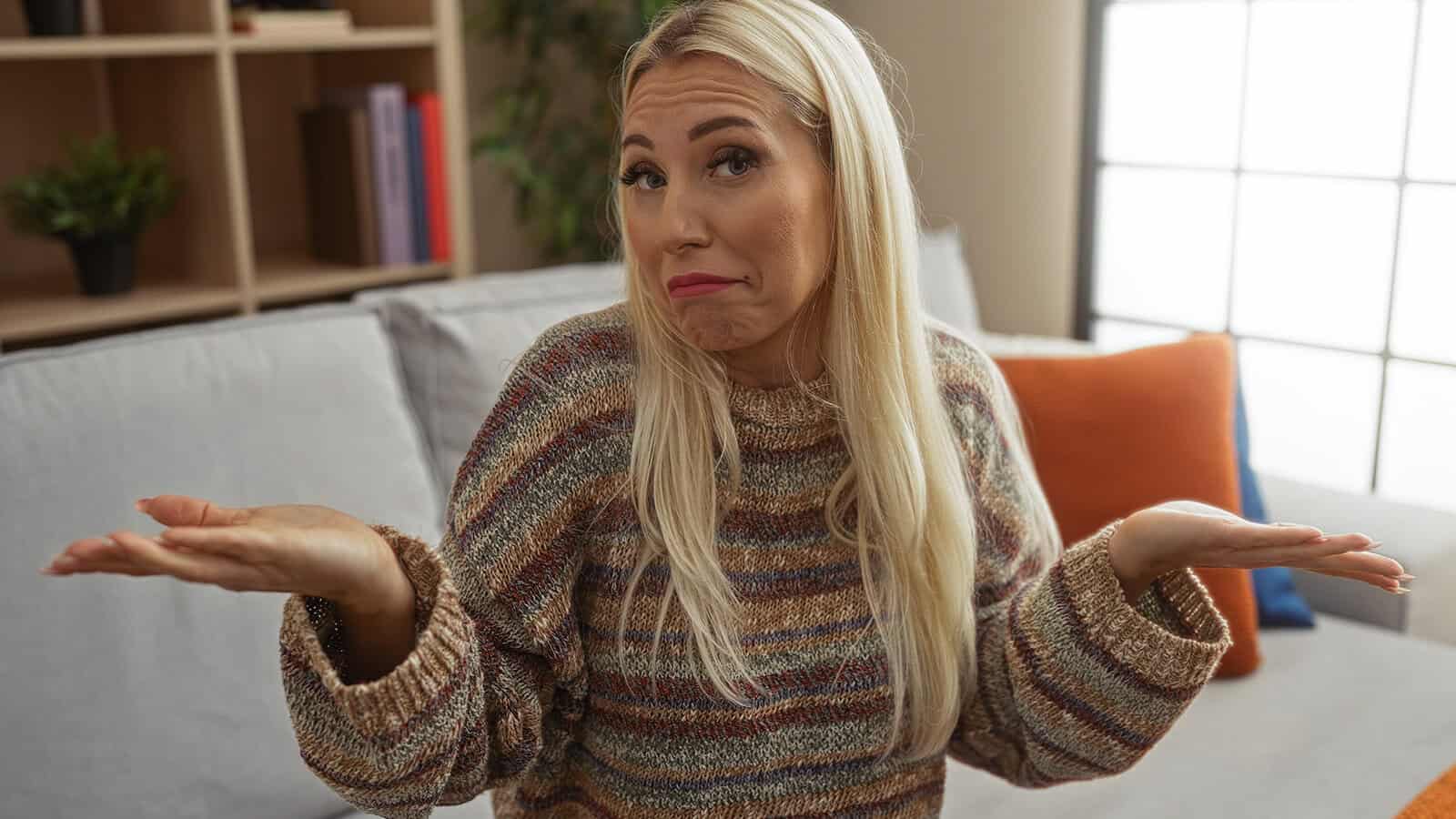 A blonde woman sitting on a sofa shrugs her shoulders with a confused expression.
