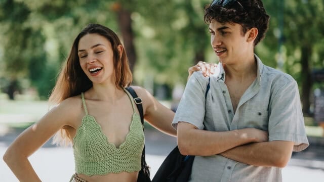 A young woman laughing as she rests her hand on the shoulder of a young man who looks at her with a smile.