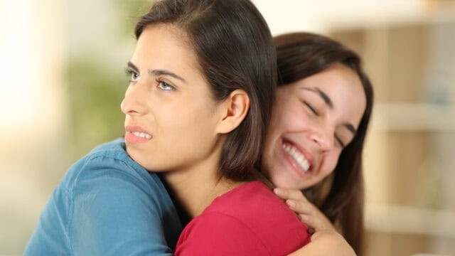 One woman smiles while hugging another woman who appears unhappy.