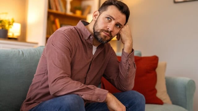 A man with a beard, wearing a button-down shirt, sitting on a sofa with his hand against his face.