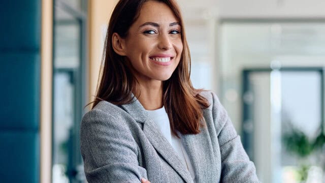 A smiling woman in a gray blazer stands in an office setting.