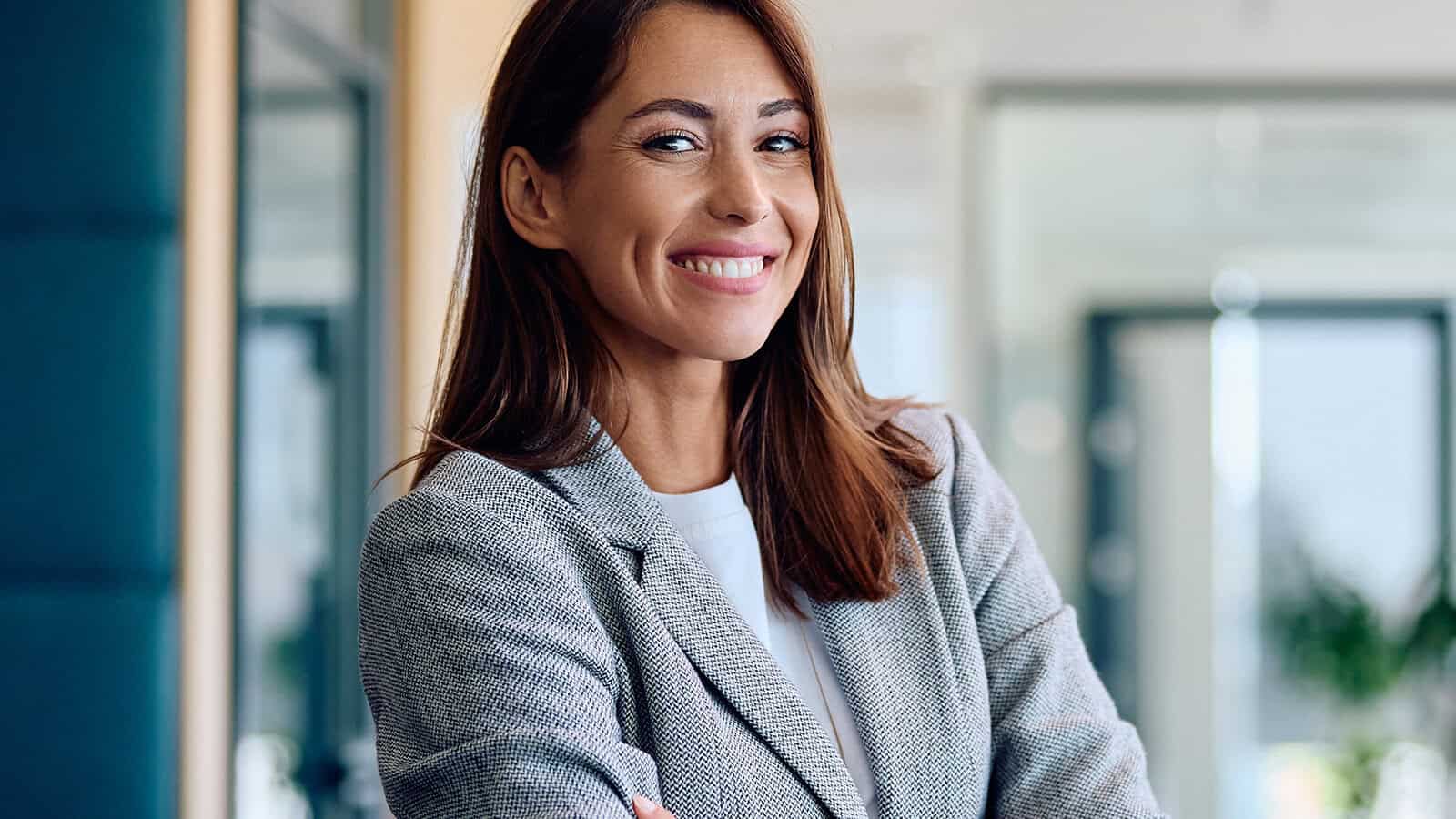 A smiling woman in a gray blazer stands in an office setting.