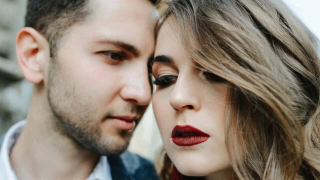A close-up of a man and a woman facing each other, both with serious expressions. The woman has bold red lipstick and styled hair, while the man has short dark hair and light stubble.