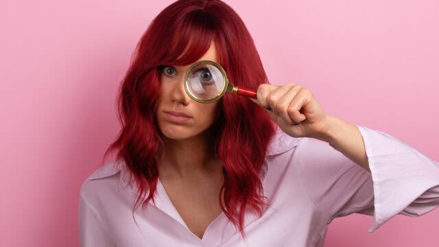 A woman with reddish hair holds a magnifying glass over one of her eyes.