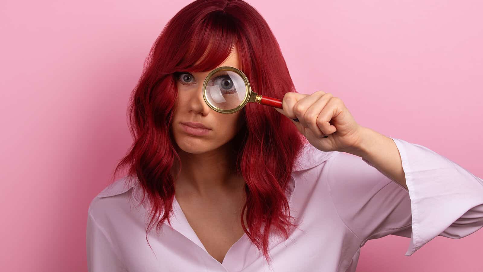 A woman with reddish hair holds a magnifying glass over one of her eyes.