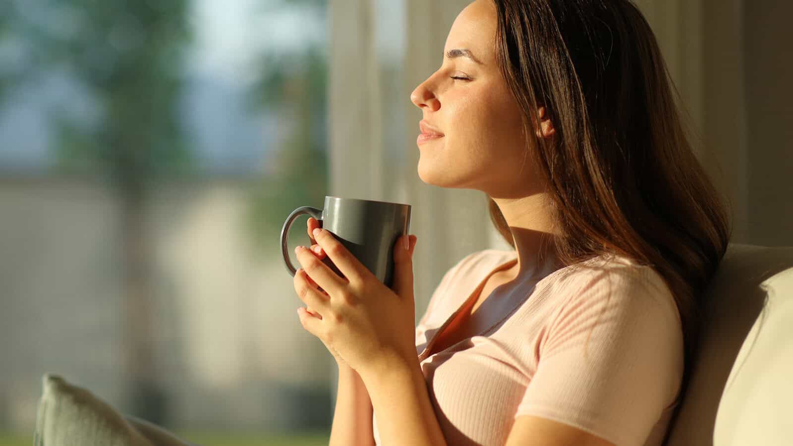 A woman with her eyes closed, smiling while holding a grey mug.