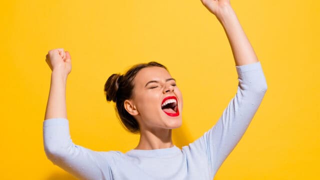 A woman cheering with her arms raised, eyes closed, and mouth open in excitement against a yellow background.