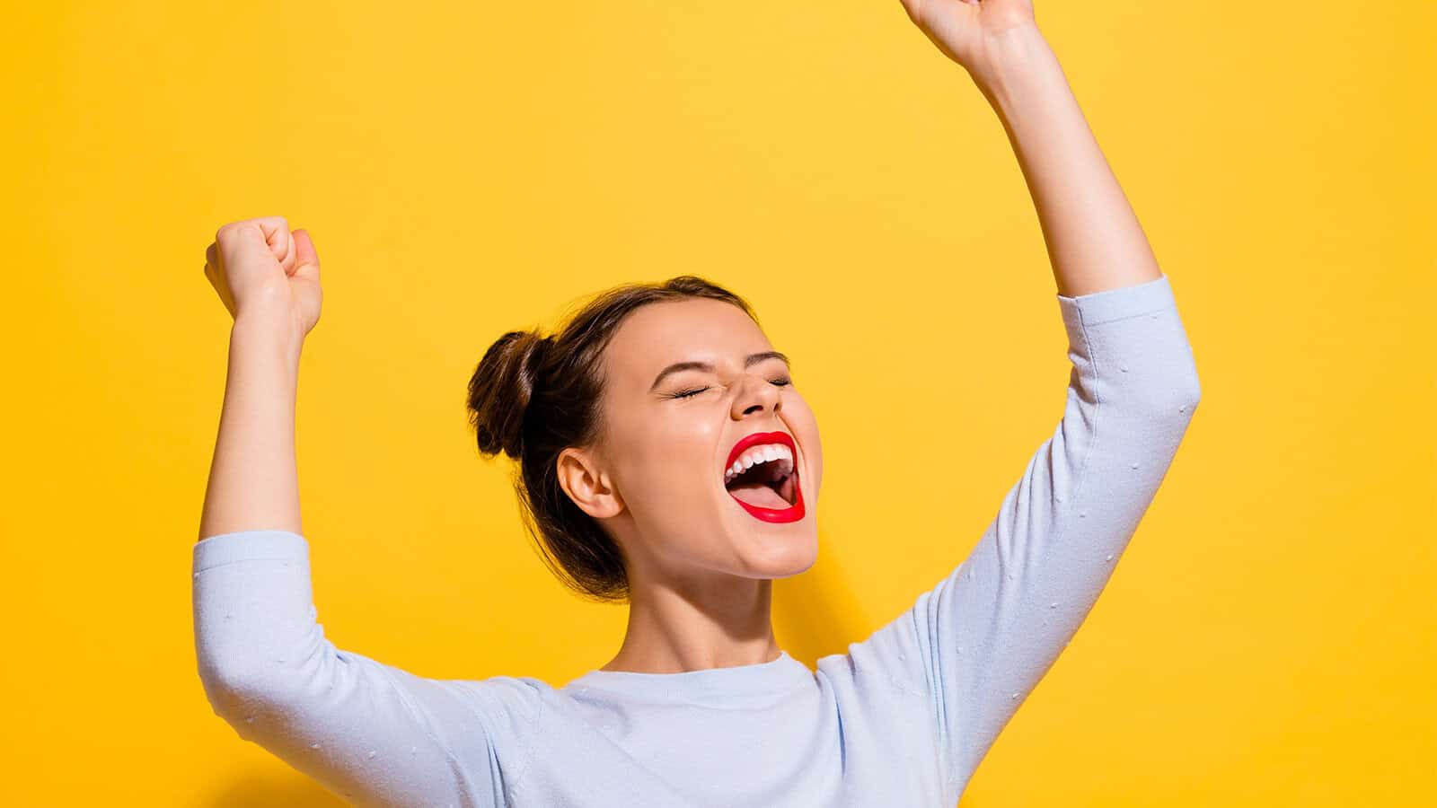 A woman cheering with her arms raised, eyes closed, and mouth open in excitement against a yellow background.