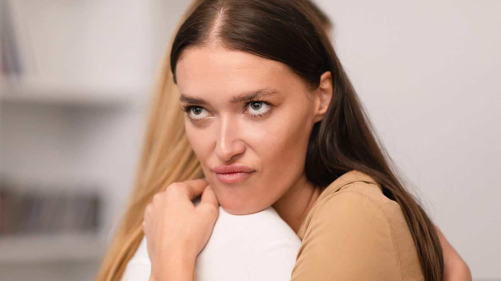 A woman with long dark hair rests her chin on the shoulder of another person while looking to the side with a skeptical expression.