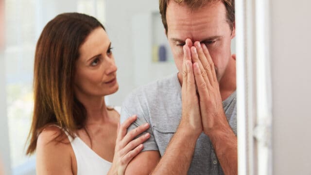 A woman places a comforting hand on the shoulder of a man who is covering his face with his hands in distress.