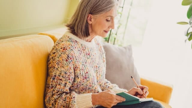 A woman with gray hair sits on a yellow sofa and writes in a small notebook with a pencil.