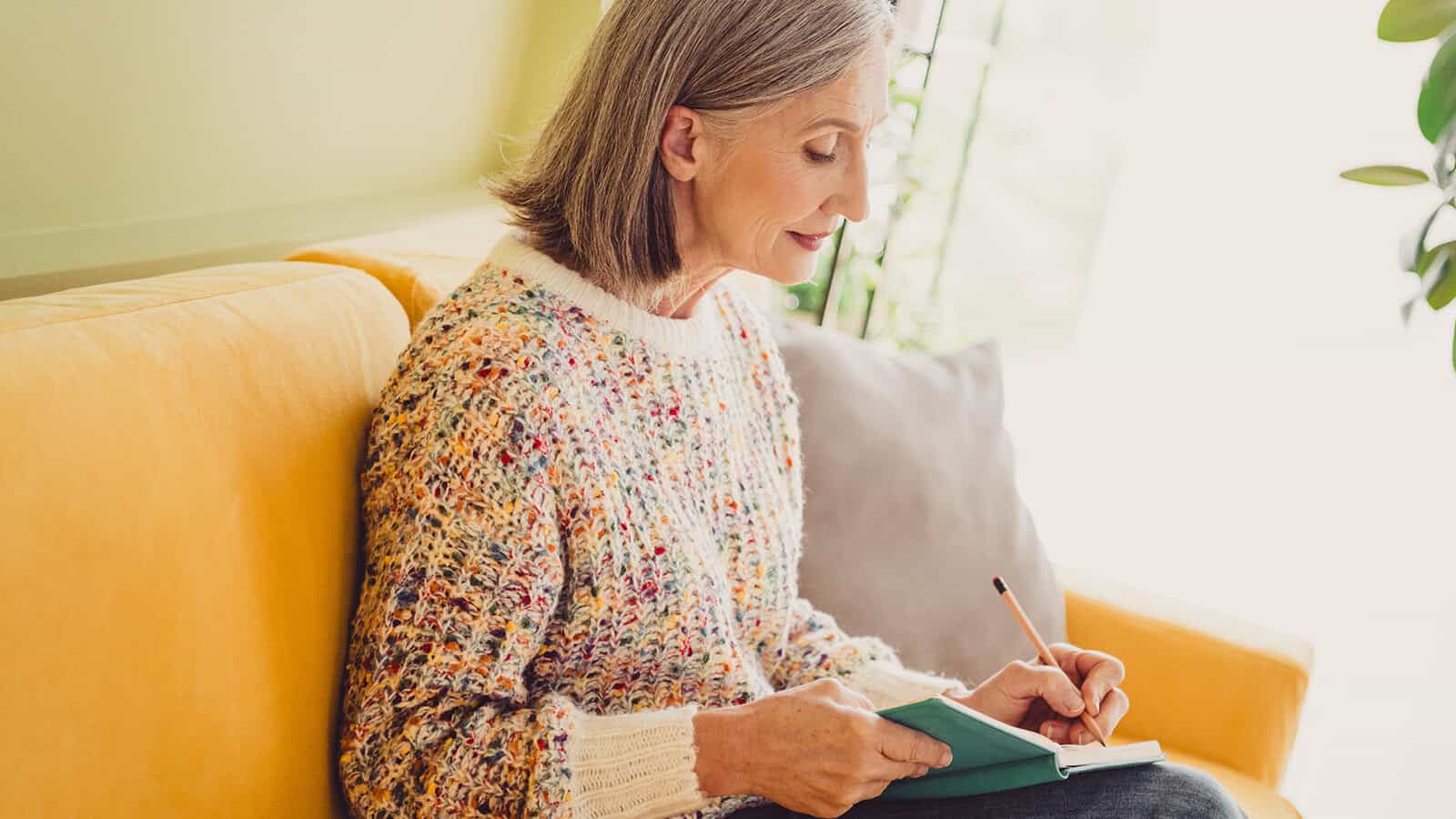 A woman with gray hair sits on a yellow sofa and writes in a small notebook with a pencil.