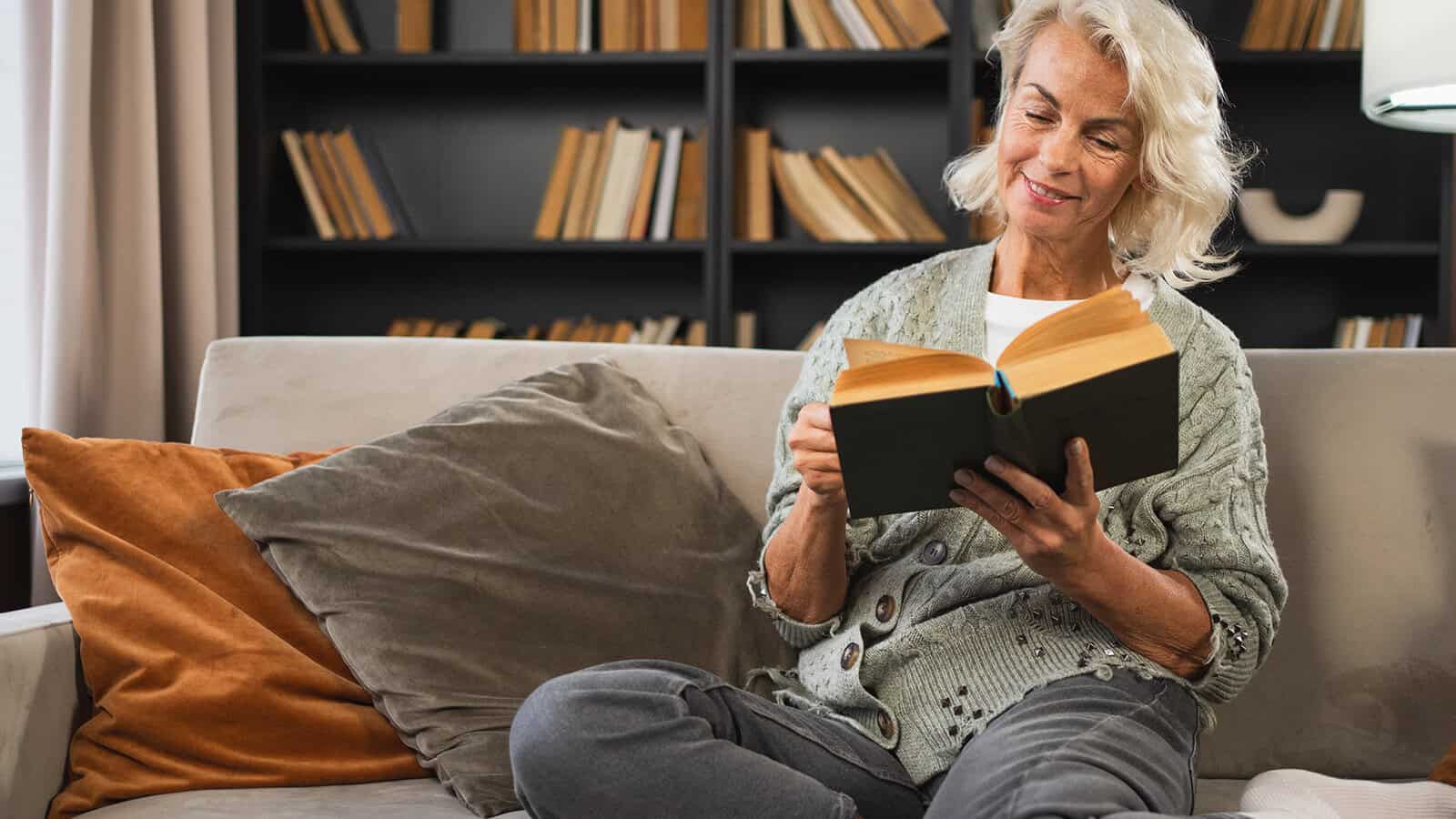 A smiling woman with blonde hair sitting on a sofa and reading an open book.
