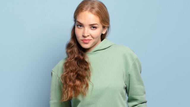 A young woman with long wavy reddish-brown hair wearing a light green hooded sweatshirt looks at the camera against a blue background.