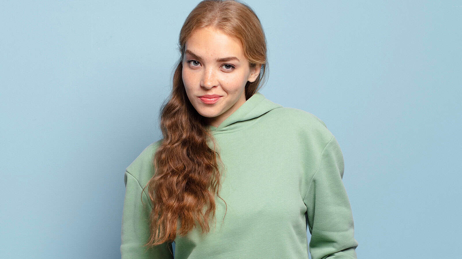 A young woman with long wavy reddish-brown hair wearing a light green hooded sweatshirt looks at the camera against a blue background.