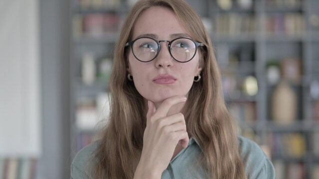 A woman with glasses looks upward thoughtfully with her hand on her chin.