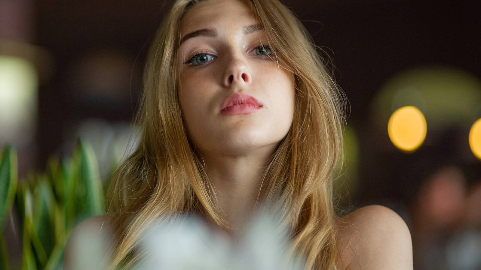 A young woman with long, wavy blonde hair looks slightly up at the camera with a neutral expression, blurred plants in the foreground, and warm lights in the dark background.