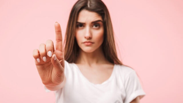 A woman with long brown hair holds up her index finger in front of her, as if signaling no or one, while looking seriously at the camera against a pink background.