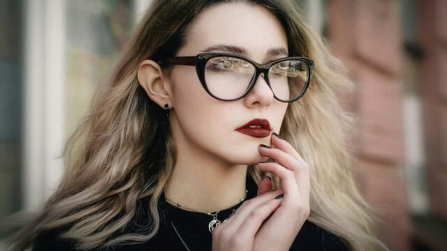 A young woman with blonde hair and glasses holds her hand to her chin.