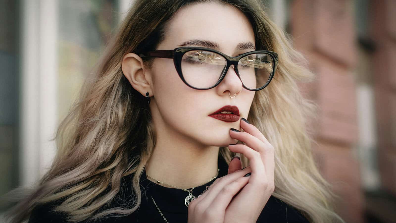 A young woman with blonde hair and glasses holds her hand to her chin.