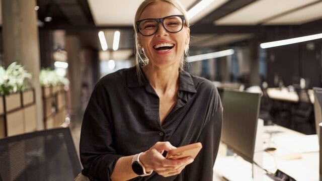 A smiling woman in glasses wearing a black shirt holds a smartphone in an office.