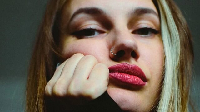 Close-up portrait of a woman with a nose ring, resting her chin on her hand and looking toward the camera.