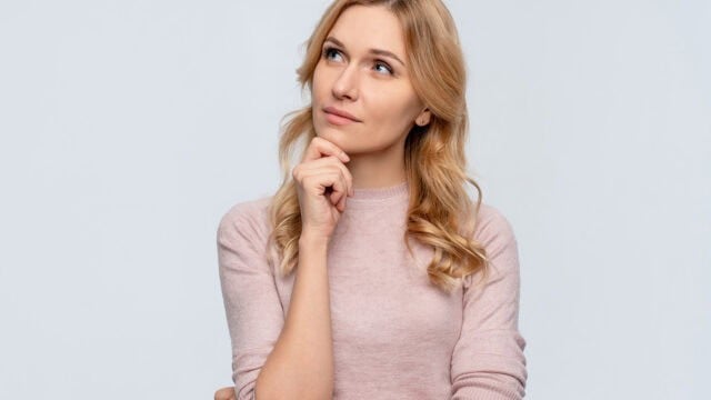 A blonde woman in a light pink sweater looking upward with her hand on her chin in a thoughtful expression.