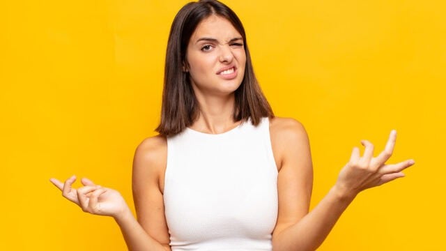 A woman in a white top wincing and making a confused, skeptical gesture with her hands against a yellow background.