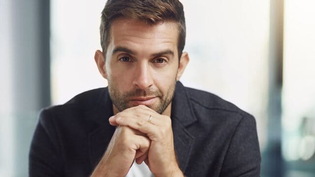 A man in a dark blazer with his hands clasped under his chin looks directly at the camera.