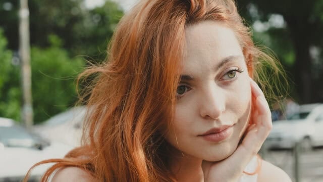 A woman with long red hair rests her chin on her hand and looks away.