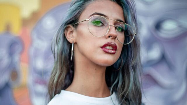 A young woman with multi-colored hair wearing large glasses and glittery lipstick.