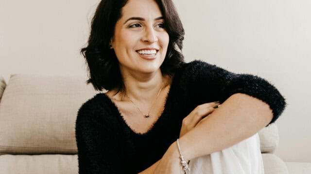A woman with dark hair and a black sweater smiling while looking off-camera.